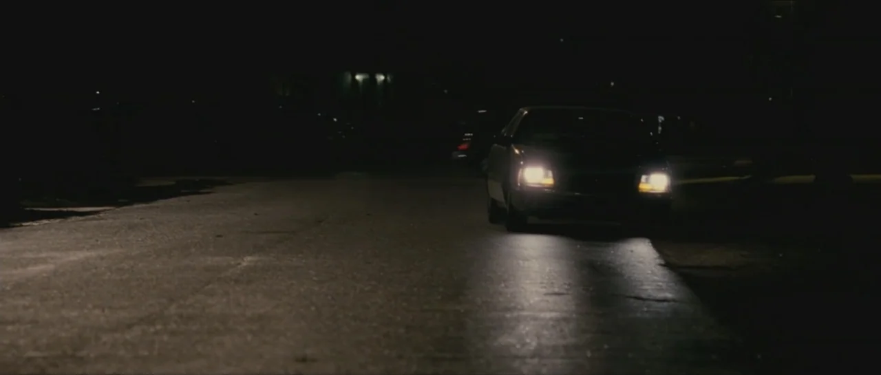 Contraband (2012) movie still: wide — A dark residential street at night features a single car parked along the curb. The headlights are…