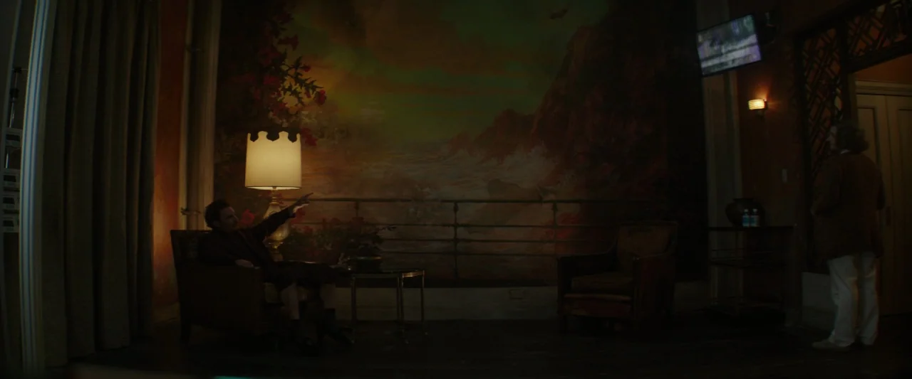 Hotel Artemis (2018) movie still: wide — A dimly lit room features a large landscape mural on the wall behind furniture. A man sits in a chair…