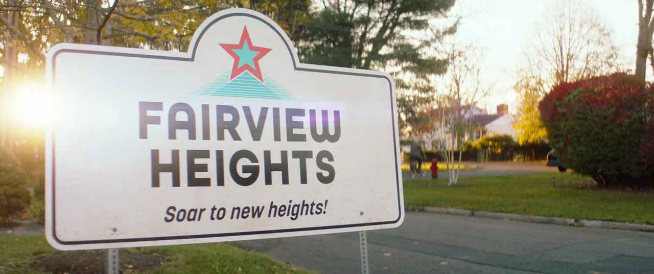 Linoleum (2022) movie still: closeup — A white sign for Fairview Heights sits in the foreground of a suburban street. Bare trees and houses line the…