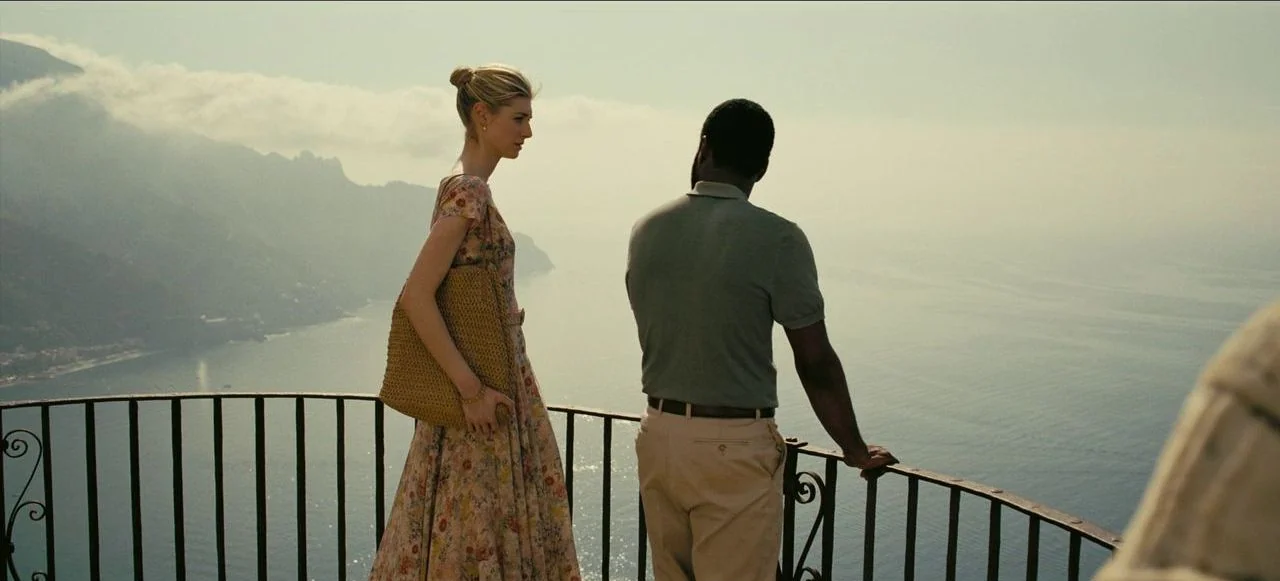 Tenet (2020) movie still: fullbody — A stone terrace with a wrought-iron railing overlooking a hazy coastal mountain range. A woman and man stand…