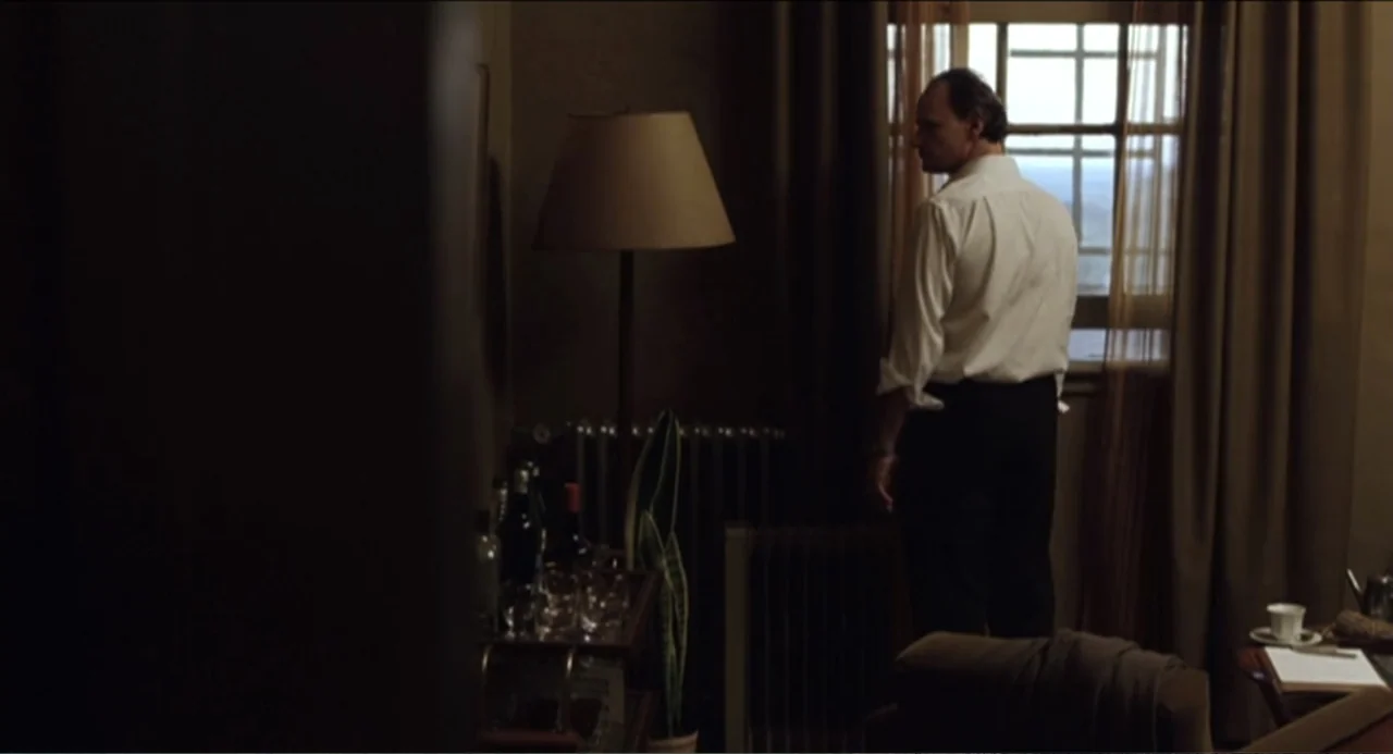 The Holy Girl (2004) movie still: medium — A dimly lit room features a wooden bar cart with glassware and a potted plant. A middle-aged man in a…