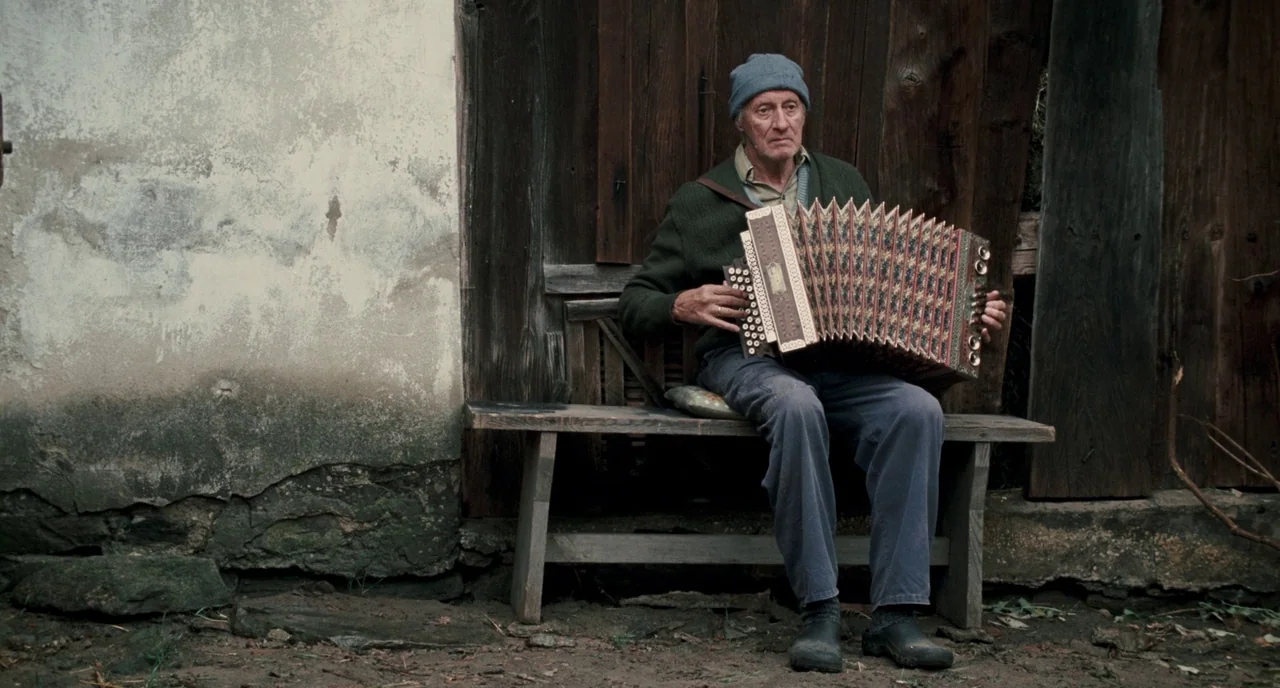 Revanche (2008) movie still: fullbody — An elderly man sits on a rustic wooden bench against a weathered barn wall. The man plays a vintage button…