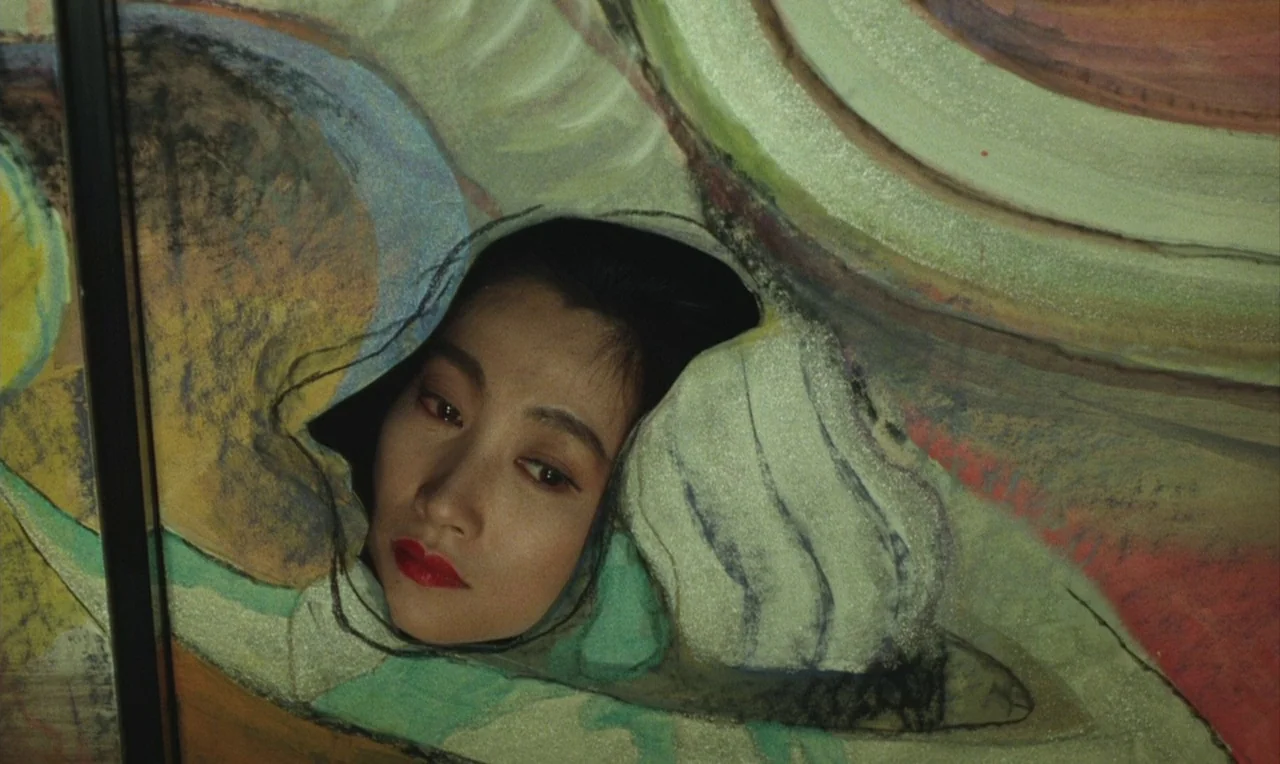 Yumeji (1991) movie still: closeup — A woman's face peers through a jagged, circular opening in a large painted canvas. The painting surface…