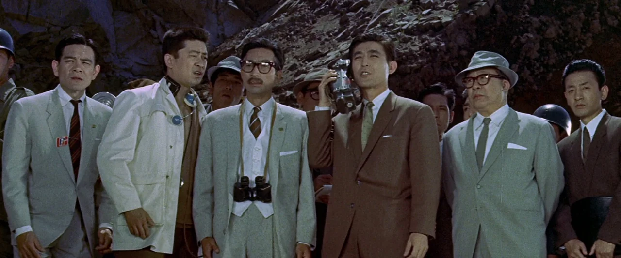 King Kong Vs. Godzilla (1962) movie still: medium — A group of men in business suits stand outdoors before a rugged rocky hillside. They look toward…