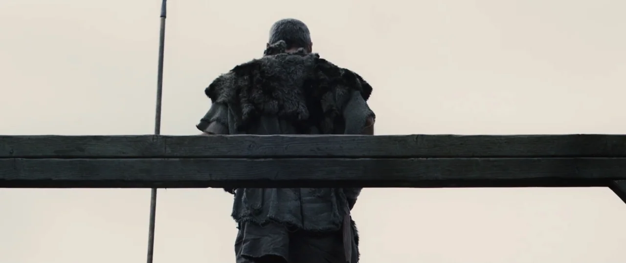 Centurion (2010) movie still: medium — A man in medieval fur-lined clothing stands behind a heavy horizontal wooden beam. He looks away from the…