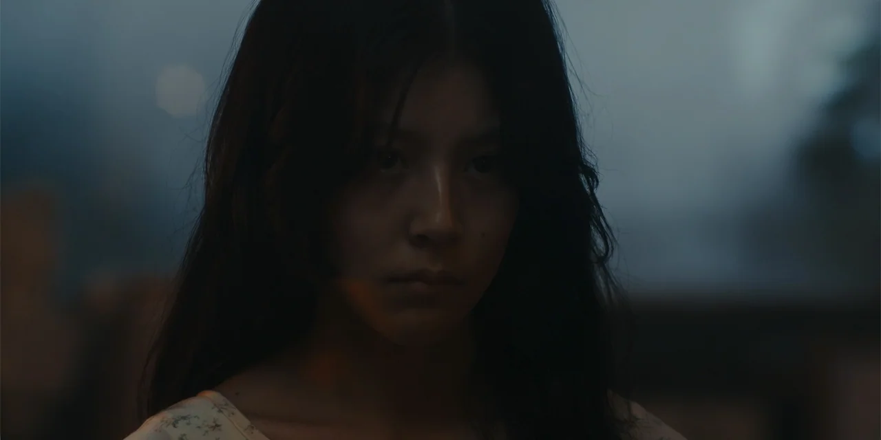Host (2020) movie still: closeup — An outdoor location at night features a dark, blurry background with subtle light sources. A young woman with…