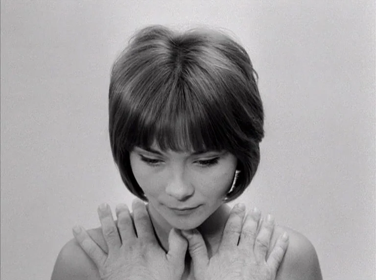 Une Femme Mariée (1964) movie still: closeup — A woman stands against a featureless light gray studio background. Hands rest on her shoulders from…