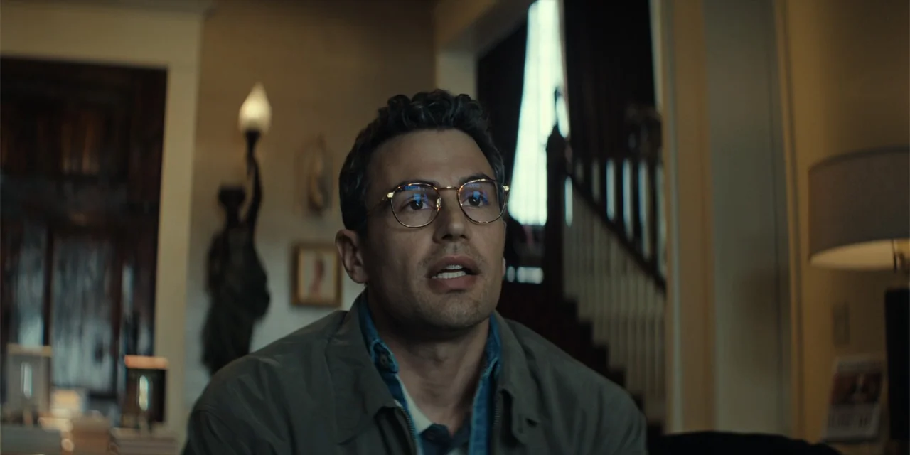 The Monkey (2025) movie still: medium — A dimly lit room features a staircase in the background and soft lamp light. A man in his thirties with…