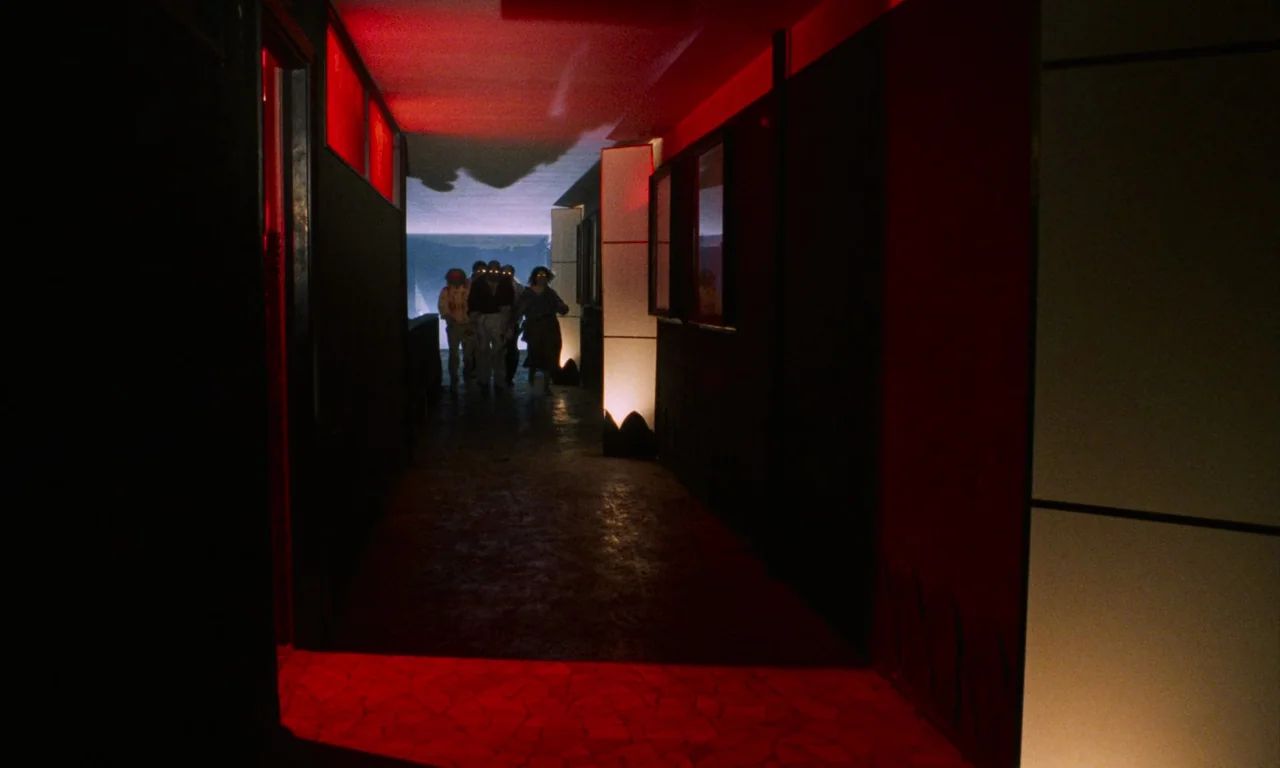 Demons (1985) movie still: wide — A long, dark corridor is illuminated by vibrant red light panels against black walls. A group of silhouettes…