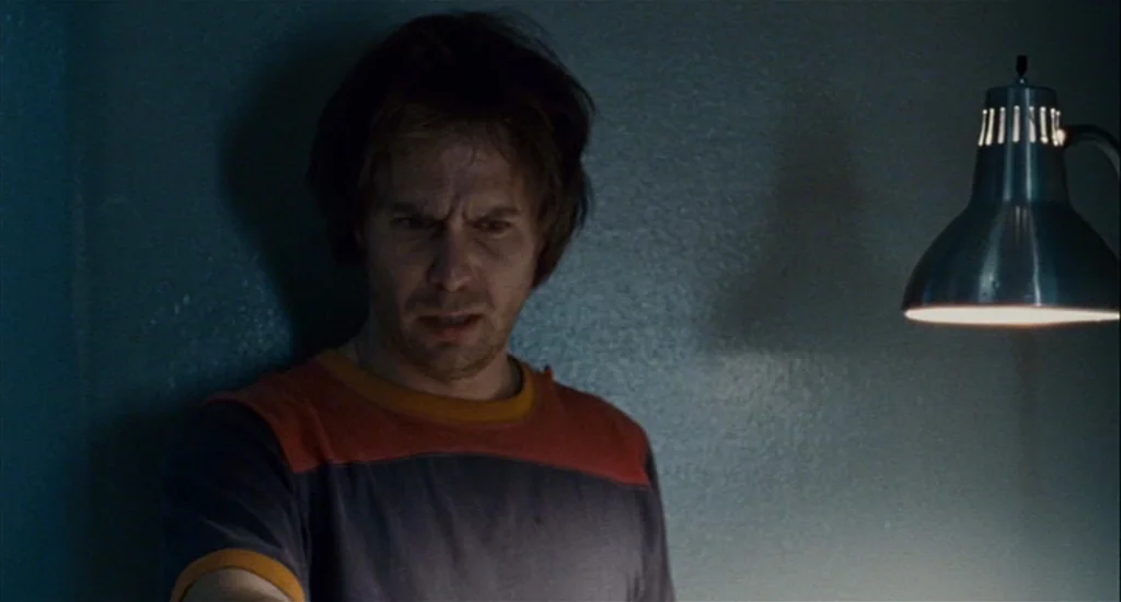 Choke (2008) movie still: medium — A dimly lit, plain interior space features a bare wall in the background. A man stands in the foreground,…