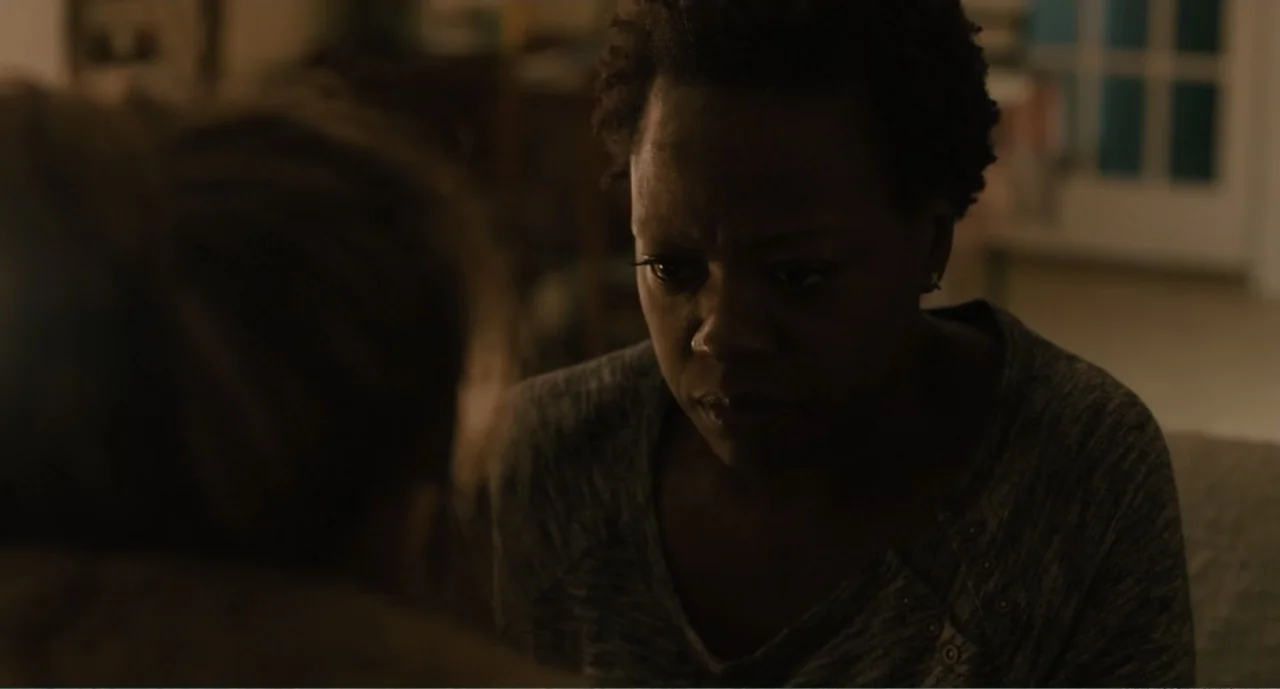 Lila & Eve (2015) movie still: medium — A dimly lit room contains two people engaged in a quiet conversation. A woman in a grey sweater sits facing…
