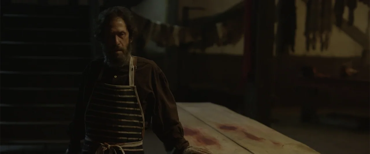 Ghosts of the Ozarks (2022) movie still: medium — A dimly lit workshop space with hanging meat and a bloodstained wooden table. A man in a striped…