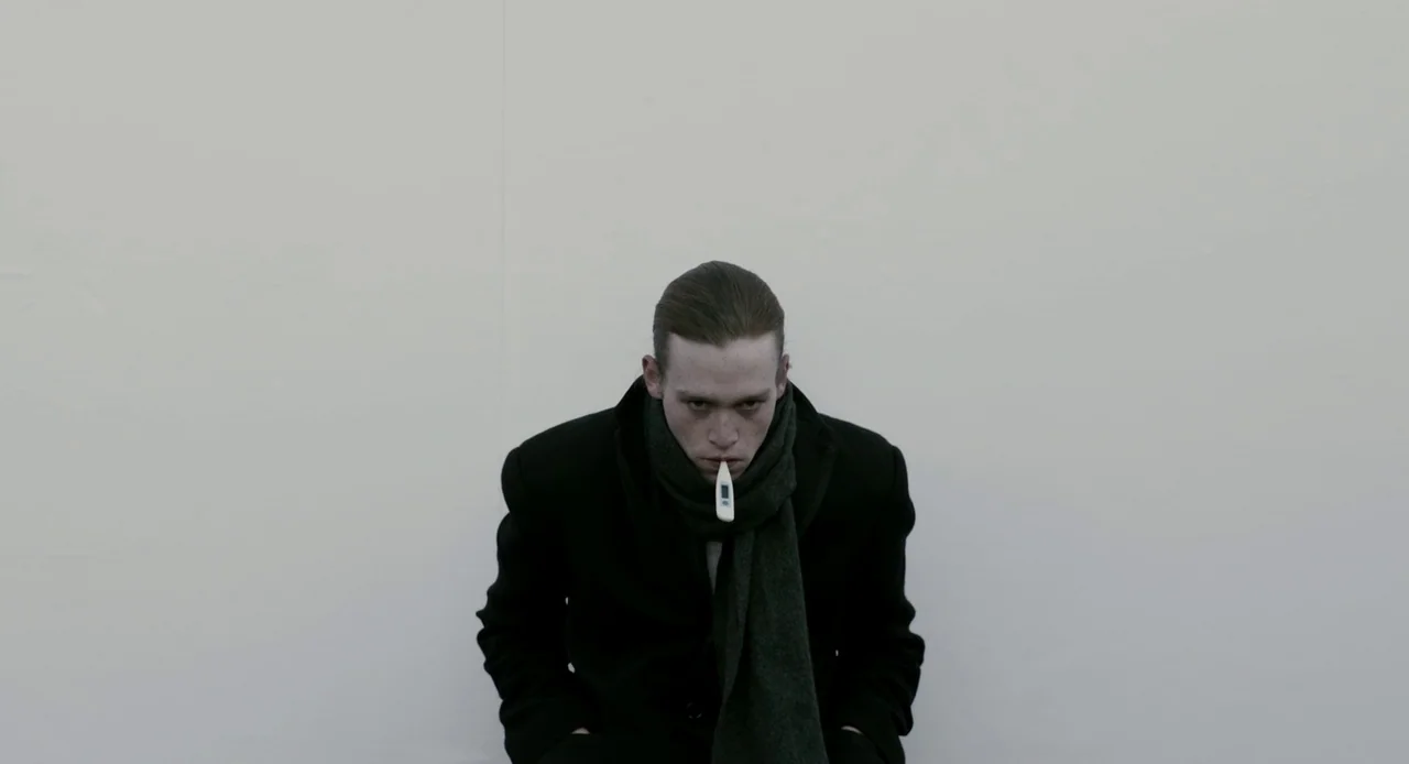 Antiviral (2012) movie still: medium — A man sits centered against a blank, off-white wall. He wears a dark coat and scarf while holding a digital…