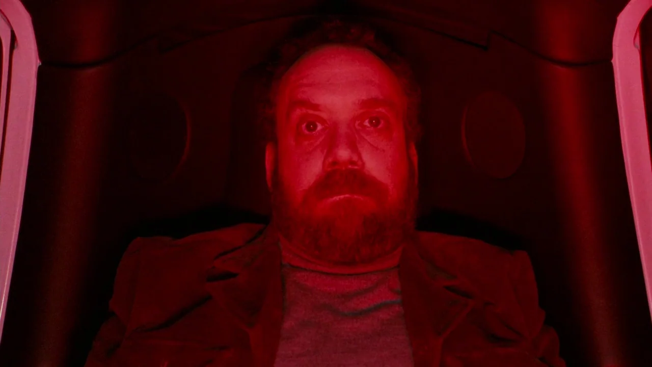 Cold Souls (2009) movie still: closeup — A man sits inside a dark enclosure lit entirely by deep red light. The man faces forward with an unblinking…