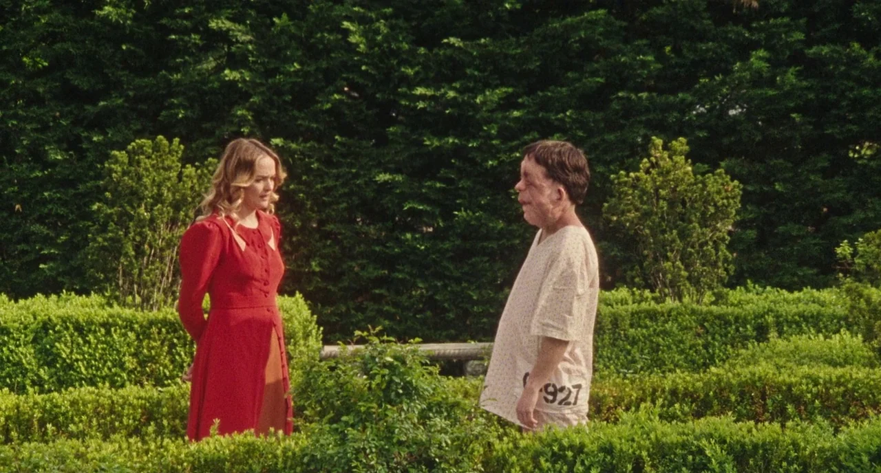 Chained For Life (2018) movie still: fullbody — A dense outdoor maze filled with manicured green hedges during the day. A woman in a red dress talks…