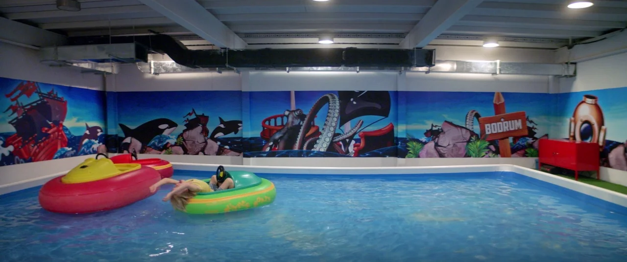 Holiday movie still: wide — An indoor rectangular pool features a painted mural with sea themes on the walls. A child drifts in a small inflatable…