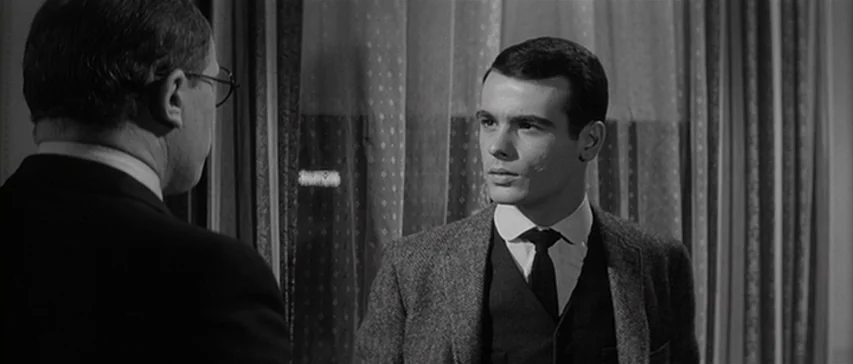 Compulsion (1959) movie still: medium — Two men stand indoors in front of a window covered by patterned drapes. A younger man faces an older man…