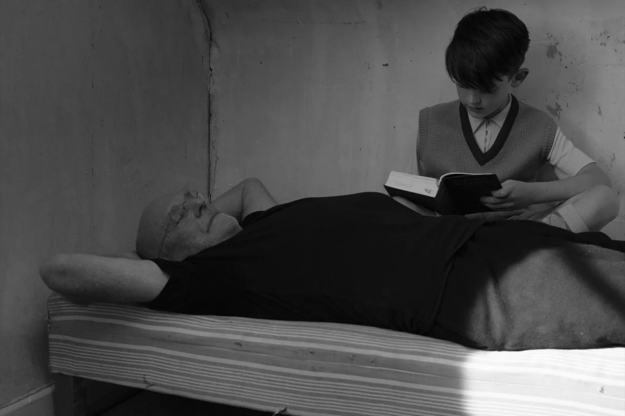 Anselm (2023) movie still: medium — A spartan room features a bed against a plain, slightly textured wall. A boy sits cross-legged on the mattress…