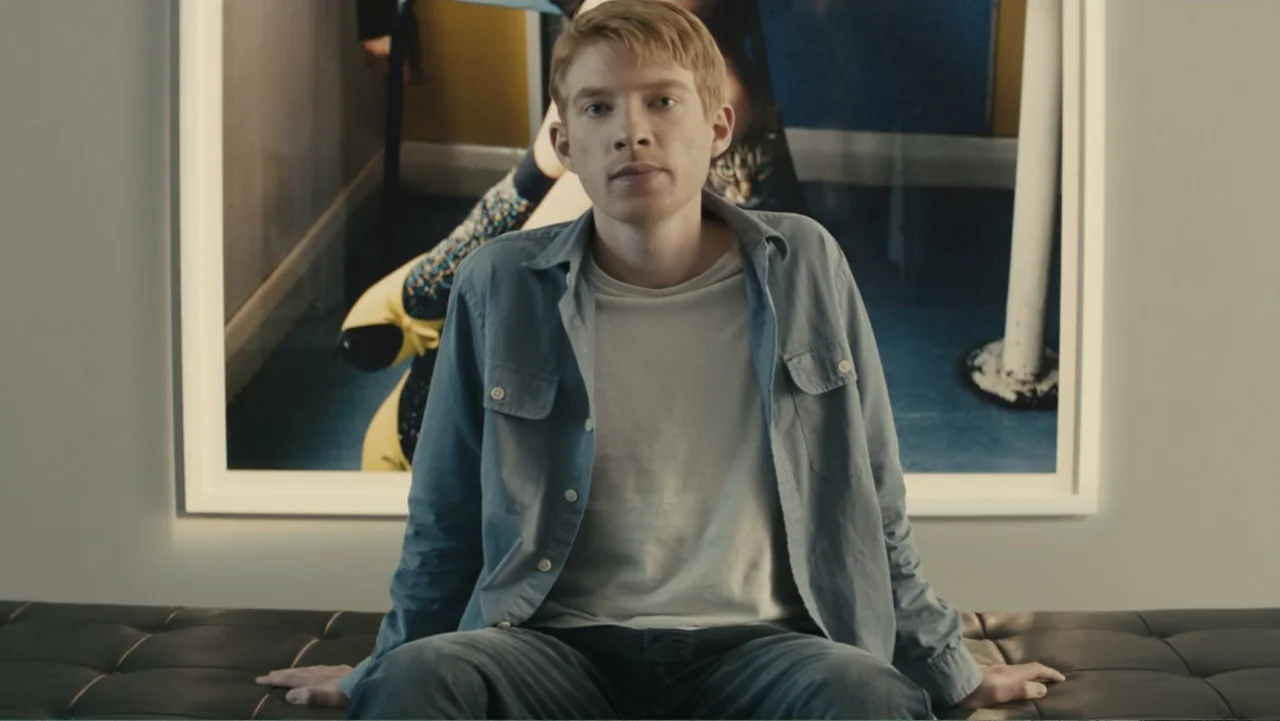 About Time (2013) movie still: medium — An interior gallery space with a large framed photograph hanging on a white wall. A young man with light red…
