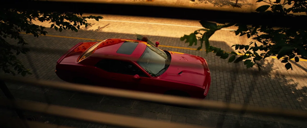 Spiral (2021) movie still: wide — A bright red coupe is parked on a sun-drenched cobblestone street during golden hour. The view is partially…