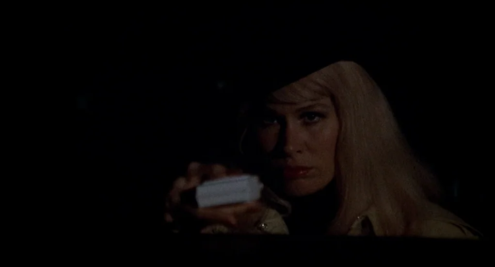 Family Plot (1976) movie still: closeup — A dark vehicle interior features a woman illuminated by low light. She extends her hand holding a small,…