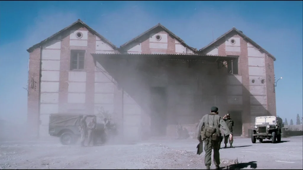 Che Part 2 (Guerrilla) (2008) movie still: wide — A large brick structure with three peaked roofs stands in a dusty, open area. Several soldiers in…