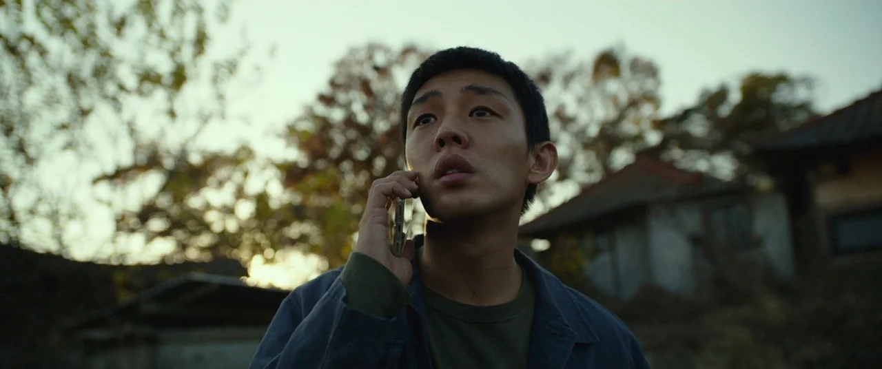 Burning (2018) movie still: closeup — A man stands outdoors in a rural area with overgrown trees and old houses. He holds a smartphone to his ear…