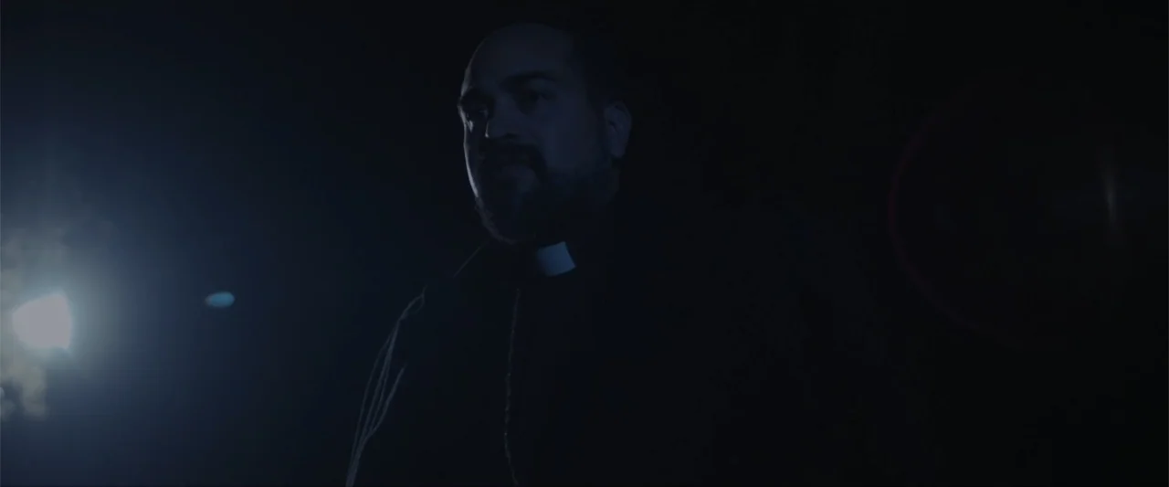 Anomaly (2006) movie still: medium — A dark outdoor setting at night with a single bright light source nearby. A man in clerical attire stands in…