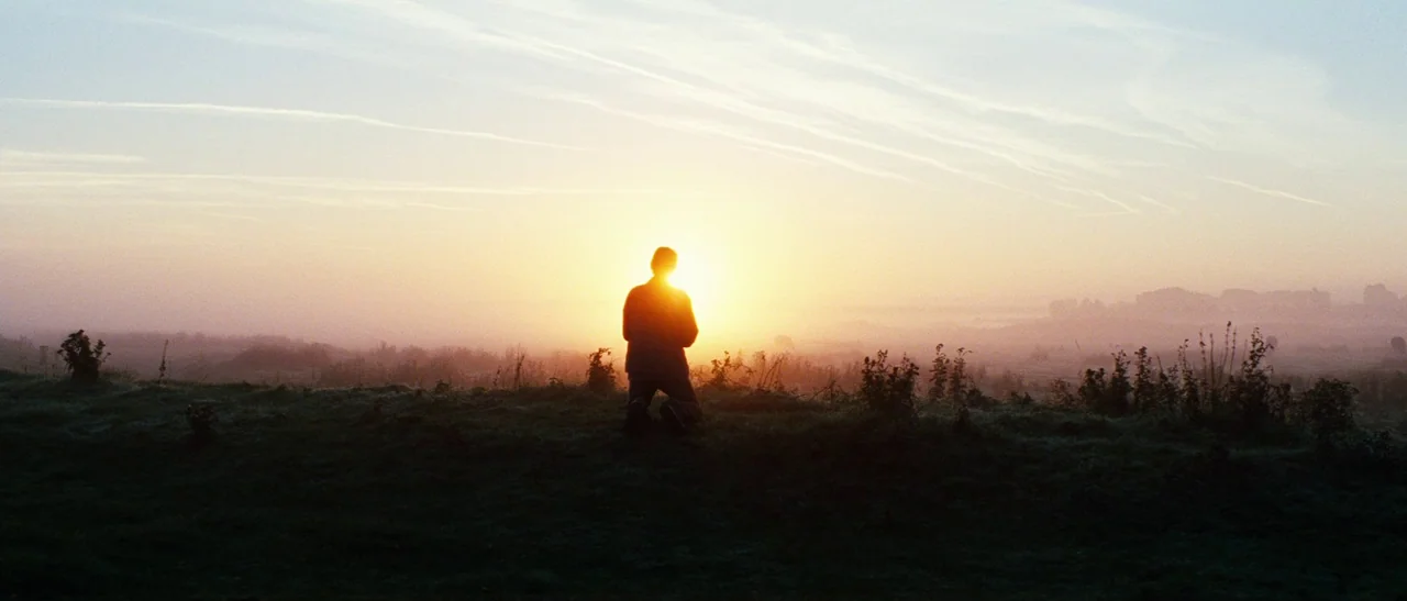 Outside Satan (2011) movie still: wide — A silhouette of a man kneels on a misty, grass-covered hill at sunrise. The bright sun sits directly behind…
