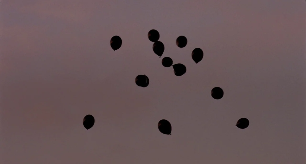 Possum (2018) movie still: establishing — A clear, muted purple evening sky serves as the backdrop for floating objects. Multiple round balloons…