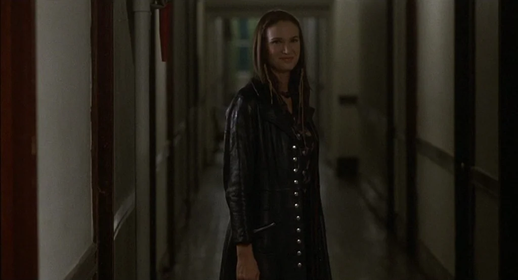 Drugstore Cowboy (1989) movie still: medium — A dimly lit, narrow hotel hallway features dark wood trim and cream-colored walls. A woman in a black…
