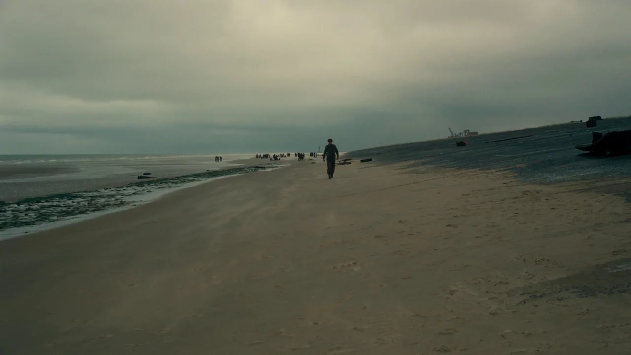 Dunkirk (2017) movie still: wide — A vast, empty beach with a cloudy sky and calm ocean under overcast weather. A lone soldier in uniform walks…