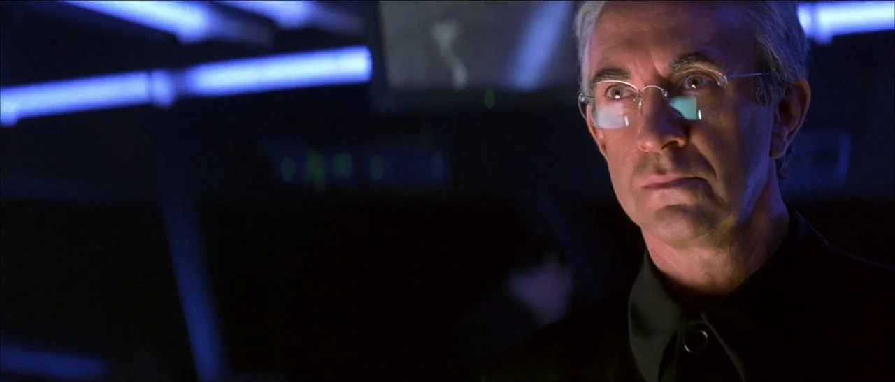 Tomorrow Never Dies (1997) movie still: closeup — A dark, dimly lit control room contains banks of monitors and blue light fixtures. An older man…