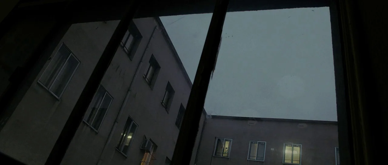 The Imposter (2012) movie still: establishing — A courtyard enclosed by multi-story institutional-style building walls is viewed from behind a dark…