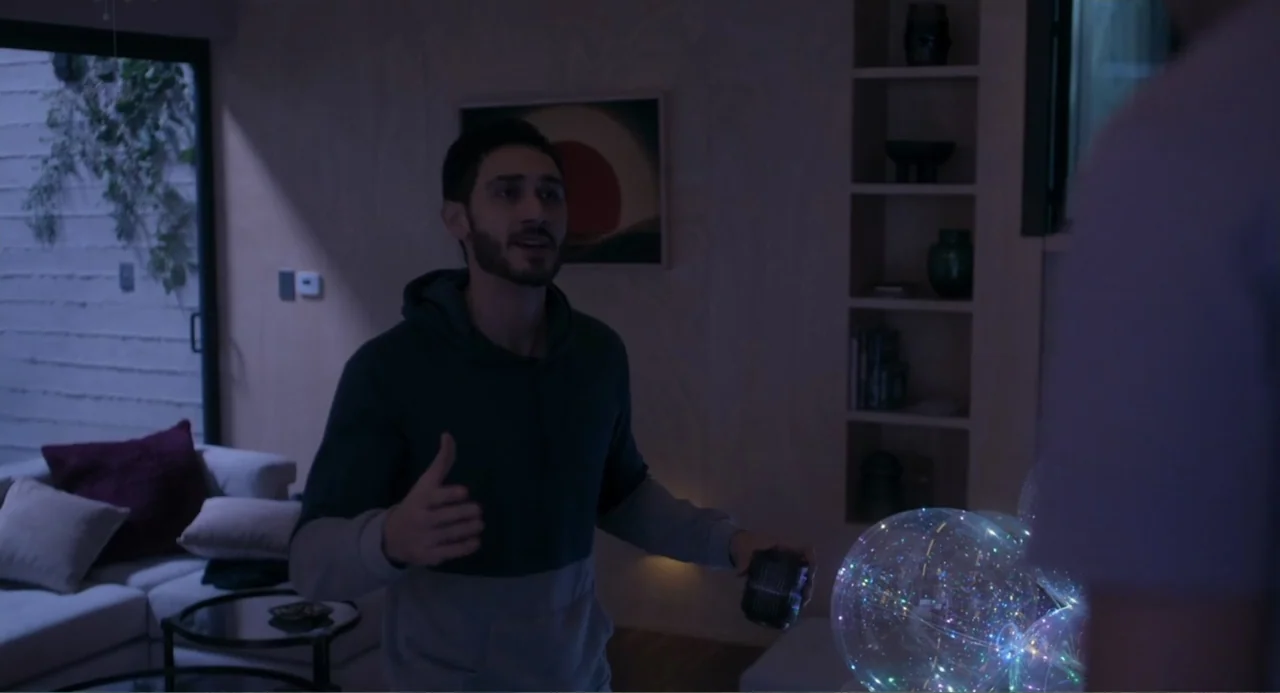 Straight (2024) movie still: medium — A modern living room at night features a couch, shelving units, and glass doors. A man in a two-toned hoodie…