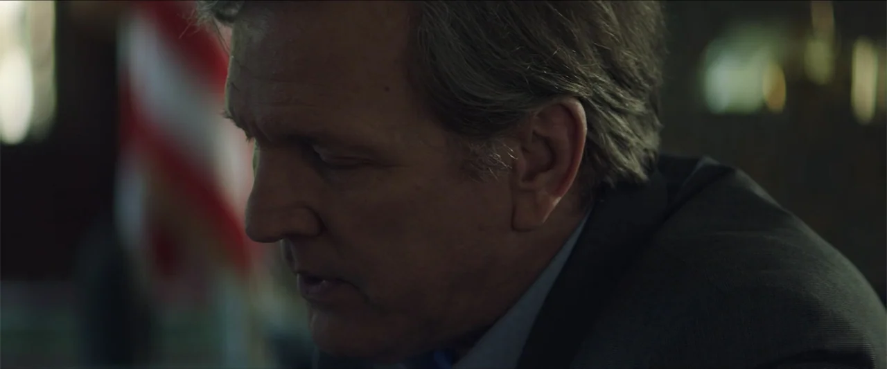 Redemption Day (2021) movie still: closeup — A dimly lit interior space features a man in professional attire seated in profile. The background…