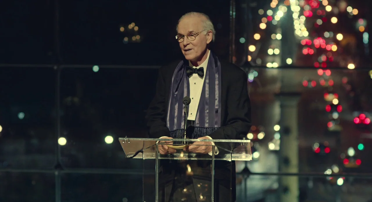 While We’re Young (2014) movie still: medium — An elderly man stands at a clear acrylic lectern in a darkened room. Bright out-of-focus city lights…