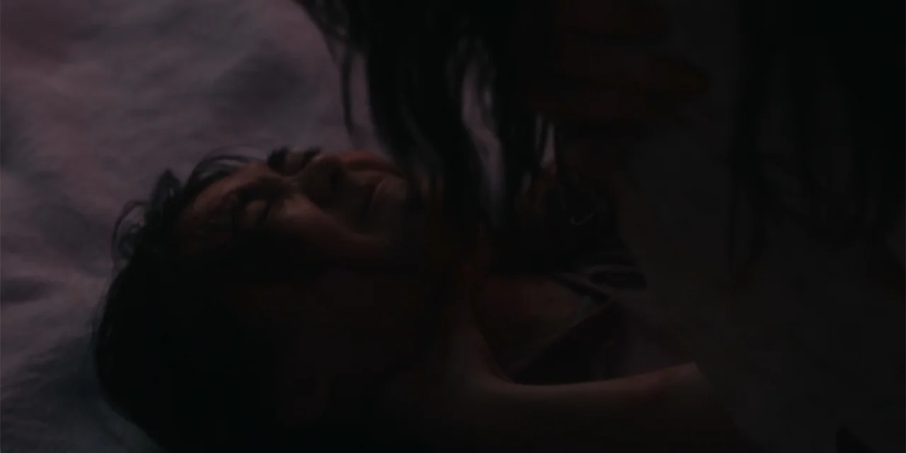 Host (2020) movie still: closeup — A dimly lit interior scene focuses on two figures in a dark setting. One person lies prone on a bed while another…