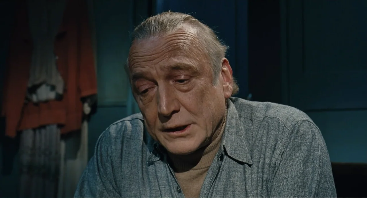 Le Havre (2011) movie still: closeup — A dimly lit indoor space with plain blue walls. An elderly man with thinning hair sits while looking downward.