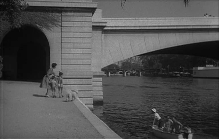 Le Signe du Lion (1962) movie still: wide — A stone bridge spans a wide river with a gravel bank in the foreground. A woman stands with two children…