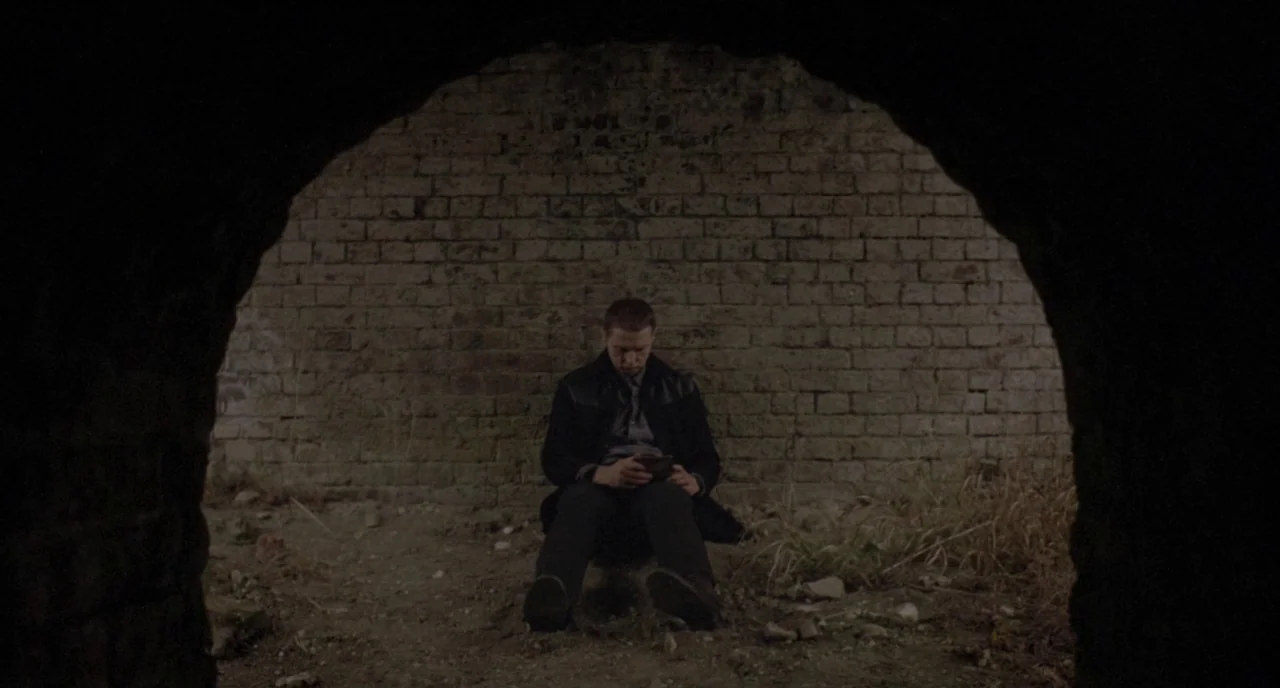 Possum (2018) movie still: wide — A man sits on the dirt floor inside a dark brick tunnel structure. He holds a small electronic device while…