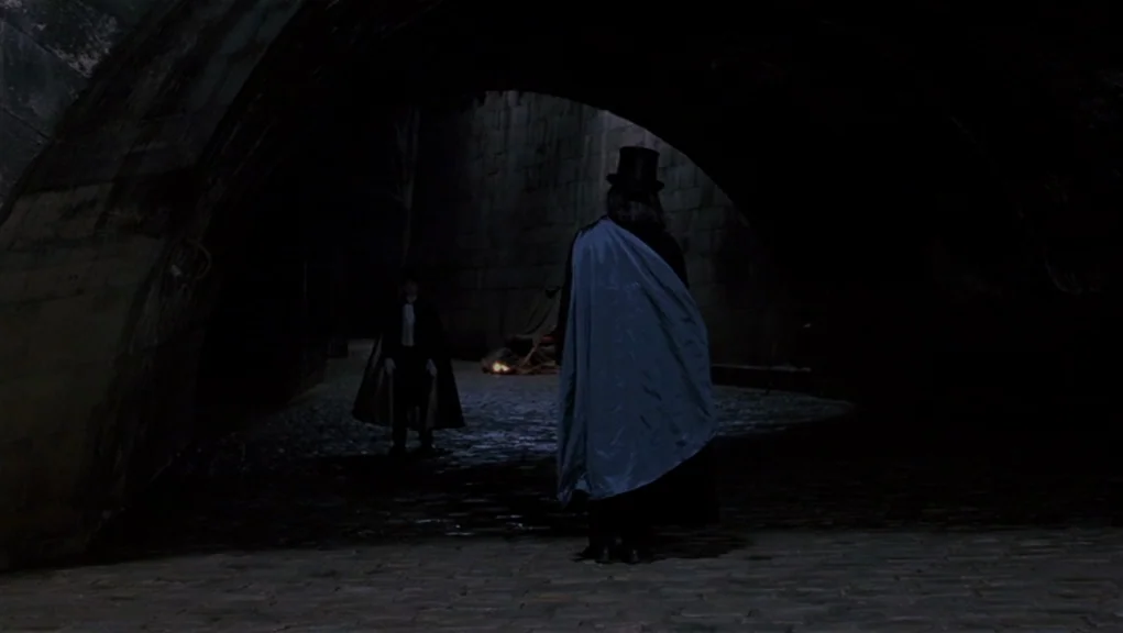 Interview With The Vampire movie still: wide — A dark stone tunnel or underpass features cobblestone ground and a low vaulted ceiling. Two men…