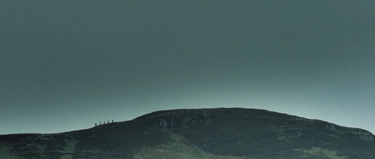 Doomsday (2008) movie still: establishing — A dark, grassy mountain ridge stretches across the lower portion of the frame. Five tiny figures walk in…