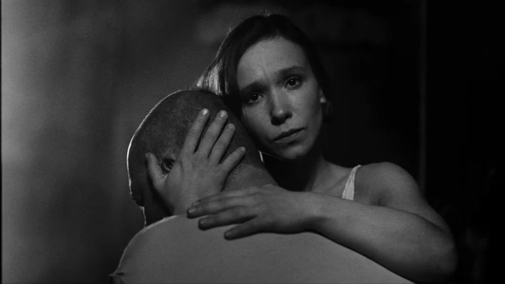 The Role (2013) movie still: medium — A dimly lit room with high contrast monochrome lighting. A woman embraces a bald man while looking towards the…