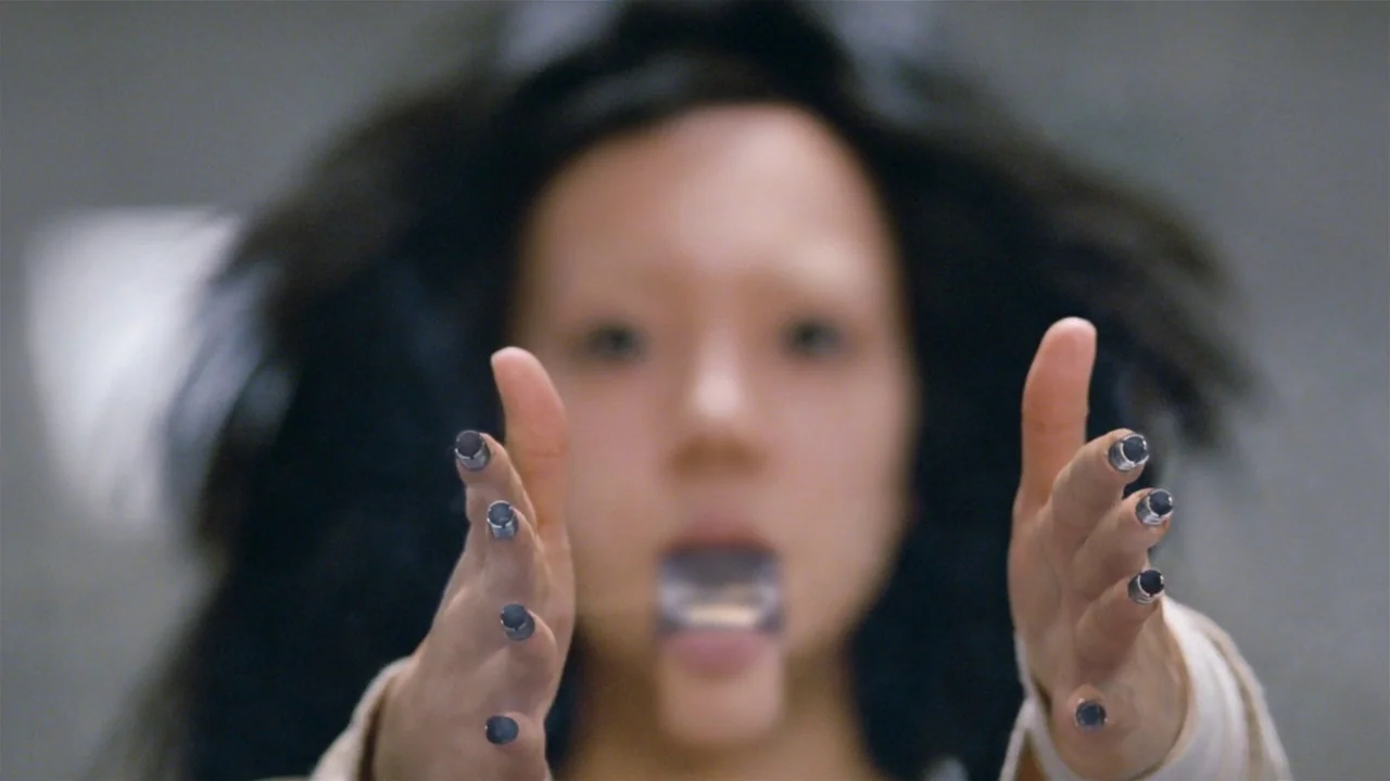 I’m A Cyborg, But That’s Okay (2006) movie still: closeup — A person with dark hair looks directly forward in a neutral room. Her hands are extended…