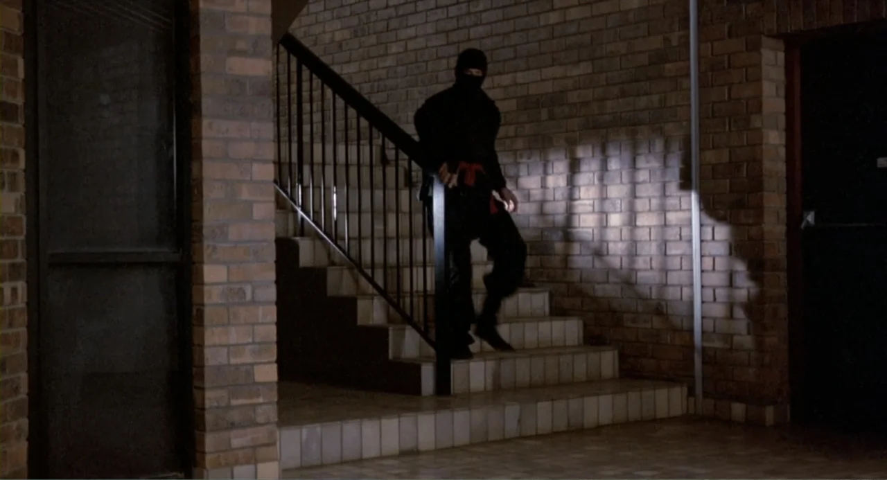 American Ninja 2: The Confrontation (1987) movie still: fullbody — A dim interior stairwell features brown brick walls and a metal handrail. A…