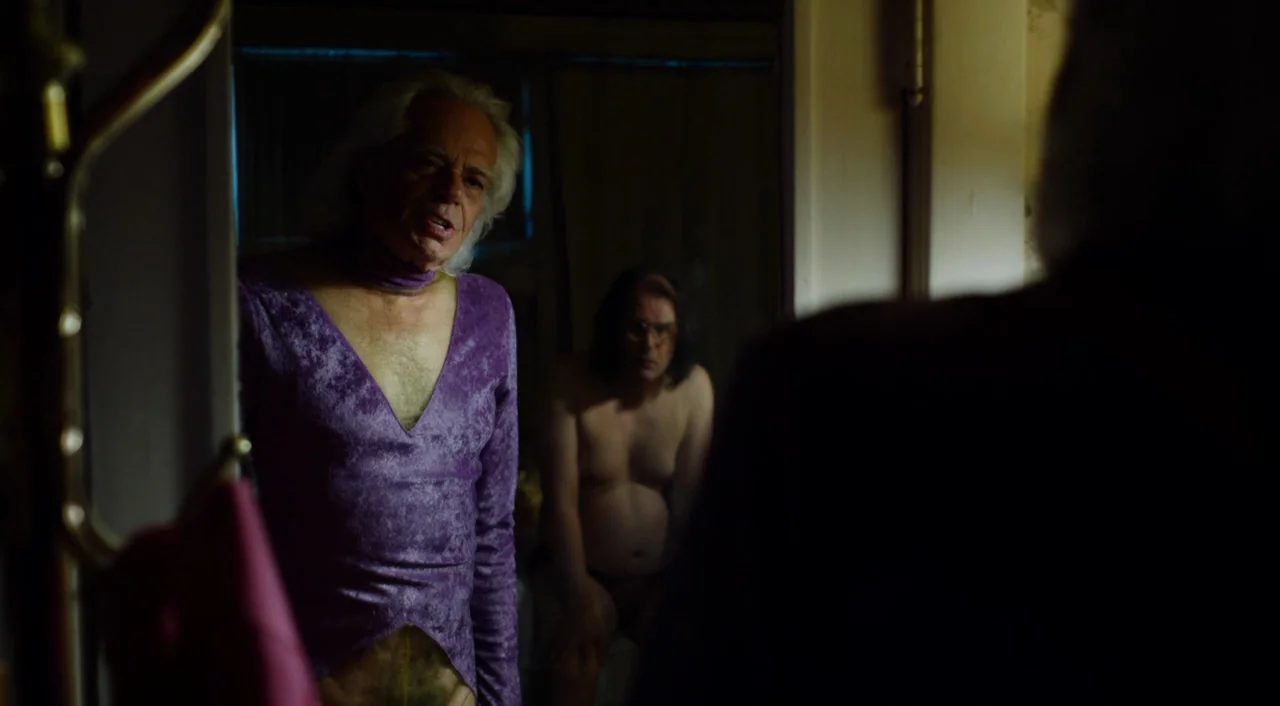 The Greasy Strangler (2016) movie still: medium — A dimly lit domestic interior space features a reflective glass surface in the foreground. An…