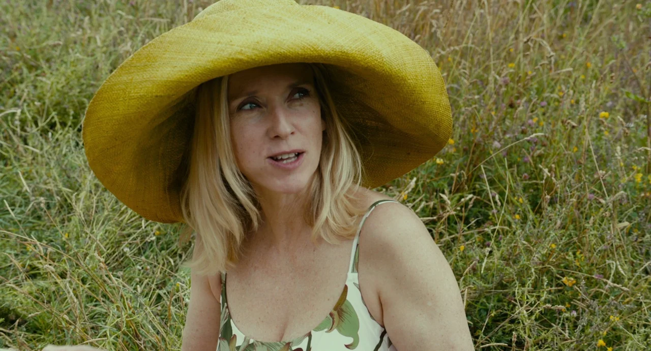 Last Summer (2023) movie still: medium — A field of tall, wild grass and scattered wildflowers surrounds a woman outdoors. The woman wears a…