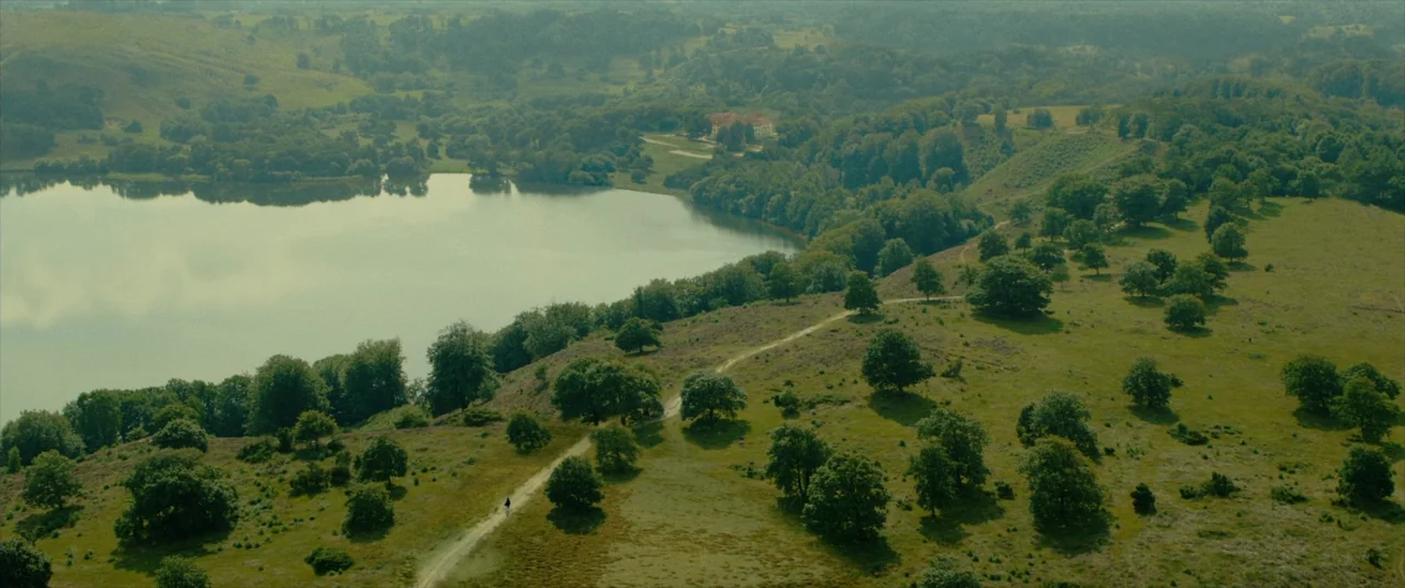 The Promised Land (2023) movie still: establishing — A vast, grassy landscape with rolling hills, a large lake, and dense woodland. A dirt path…