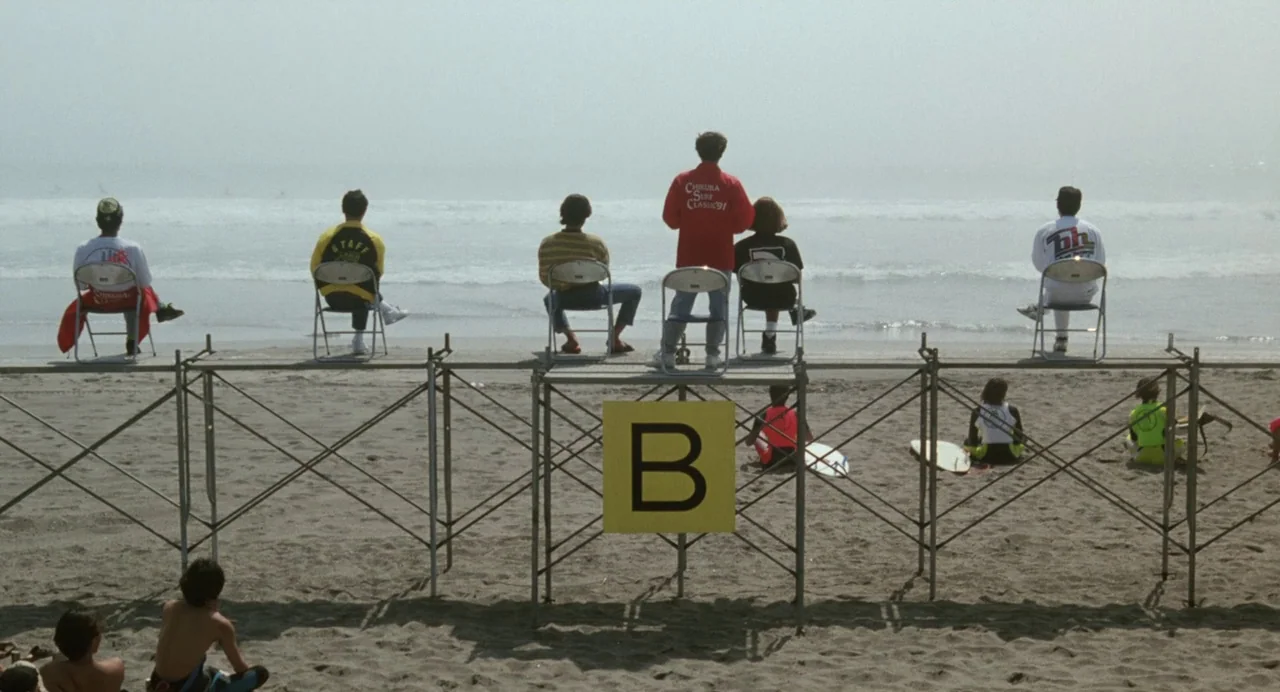 A Scene at the Sea (1991) movie still: wide — A group of people sit on raised metal scaffolding overlooking a foggy ocean beach. A large yellow sign…